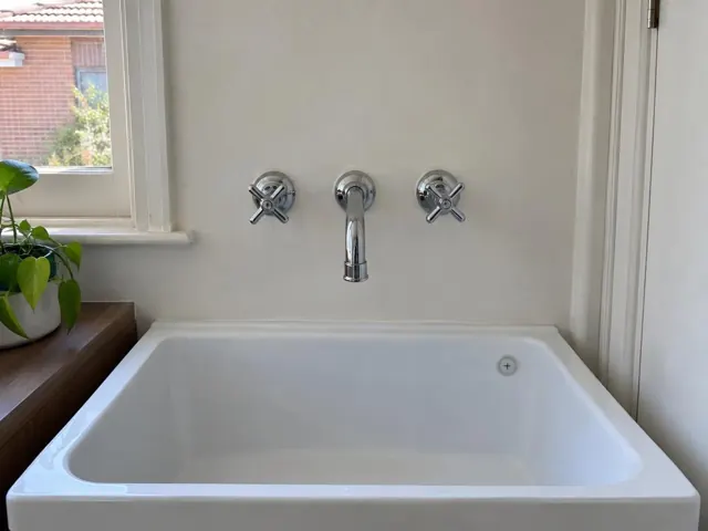 Wall-mounted tap above a white laundry tub with a dry sink and timber bench in a home interior in Coogee
